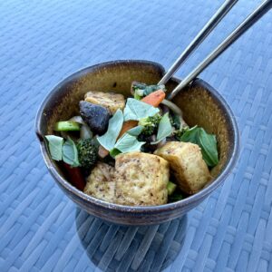 A bowl of vegan stir fry with air-fried tofu, shiitake mushrooms, broccoli, bell peppers, and fresh basil, captured on a woven glass table with chopsticks resting on the bowl’s rim.