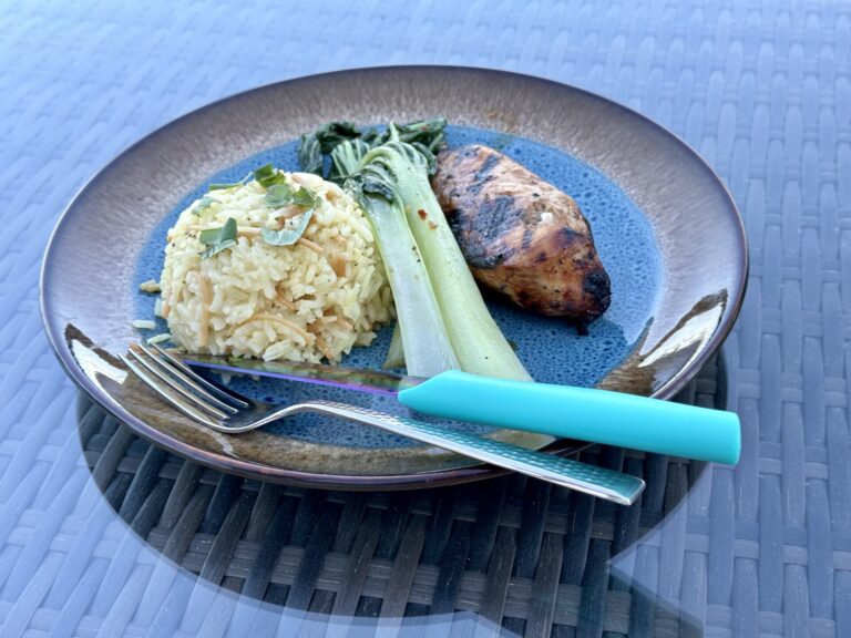 A plate featuring a grilled chicken breast, a serving of rice pilaf, and a side of sautéed bok choy. The dish is presented on a blue and brown ceramic plate with a teal-handled fork resting on the side.